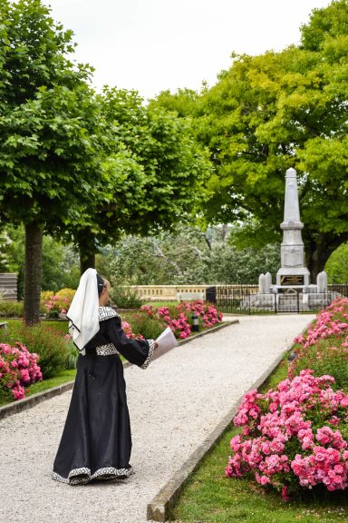 Femme en costume traditionnel marchant sur un chemin fleuri près d'un monument.